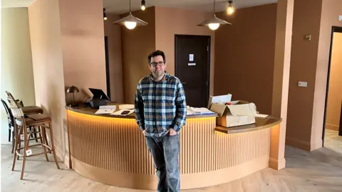 A man stands, with his hands in his pockets in front of a curved wooden reception desk. The floor and all the walls are wood-lad. Behind him is a toilet door and to his side are several chairs. 