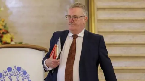 Media PA Mike Nesbitt berjalan keluar dari perakitan Irlandia Utara dengan file di bawah lengan. Dia memiliki rambut garam dan merica dan kacamata berbingkai hitam dan mengenakan jaket jas gelap, kemeja putih dan dasi merah.