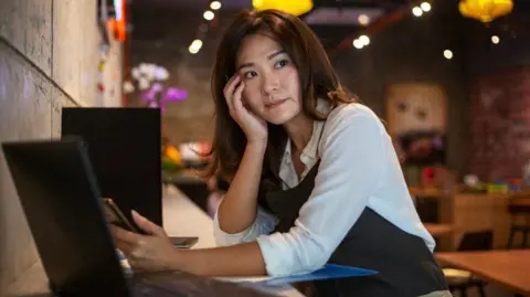 Getty Images A young Asian woman wearing an apron looking concerned at a laptop screen with a bar restaurant behind her