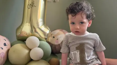 Dave Smith Rupert has brown curly hair and is wearing in a one piece brown-and-white striped top with a brown number one it and a large gold balloon and stuffed animals beside him