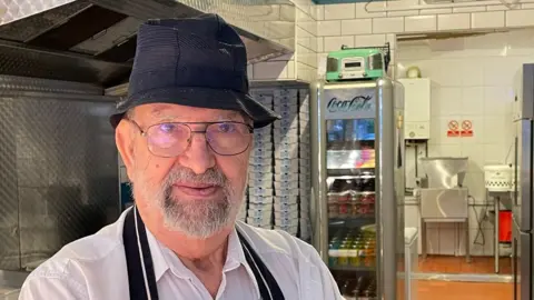 Annabel Amos/BBC John Panayis stands in front of frying equipment in a white shirt, black apron and black hat.