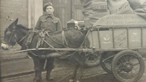 Soldiers of Shropshire Museum A black and white image of a donkey hauling a cart. The cart is full of sacks of letters. A man in an army uniform is standing behind the donkey.