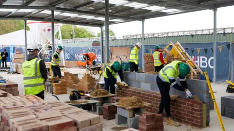 NHBC General view of apprentices in protective gear practicing bricklaying. They are outside in a large yard. It has a metal roof held up by metal poles.