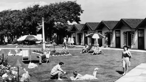 Getty Images A black and white photo showing men and women sitting and lounging around a grass area, with holiday cabins in the background.