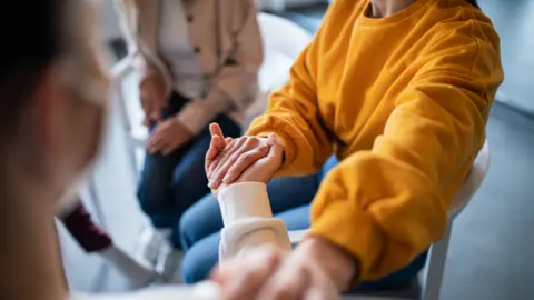 GETTY A person in a white jumper holds the hand of a person in an orange jumper whilst they put another hand on their shoulder. They are sat in a circle on white plastic chairs in some type of support group.