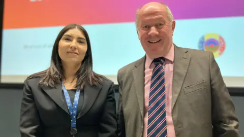 Jon Wright/BBC Simona Lazar and Tim Passmore stand next to each other smiling, there is a lecture theatre screen behind them. Ms Lazar is wearing a black jacket and blue lanyard. Mr Passmore is wearing a grey jacket with a pink shirt and striped tie. 