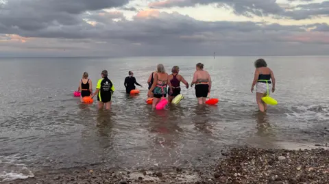 Amanda White Eight women in swimming costumes and wearing fluorescent buoyancy aids walk into the sea for a swim and a natter.