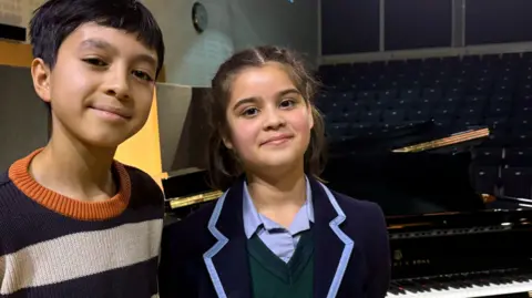 Two children smile into the camera, one is wearing a stripey jumper and the other is wearing a school uniform.