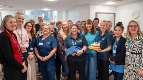 West Suffolk NHS Foundation Trust Kate Turner smiles at the camera surrounded by her colleagues. She wears a navy cover over her head, as well as a navy polo top with the West Suffolk NHS Foundation Trust logo on it. She also wears red glasses with a lanyard around her neck.