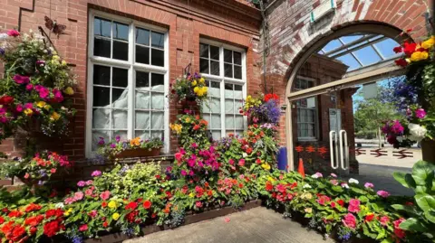 Sarah-May Buccieri/BBC Red, pink, yellow and purple flowers in flower beds and in hanging baskets surround windows and an arched doorway at Bridlington station