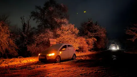 A car parked towards the side of the road covered in snow. The light is reflecting an orange colour.