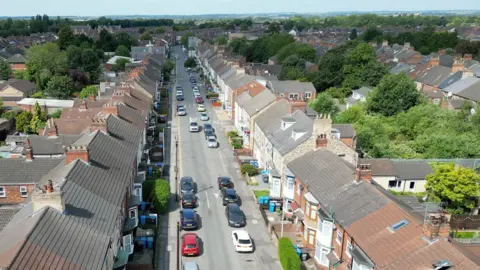 An aerial view of roof tops, and on the ground blue and black bins lining a street of terraced housing. The streets has a mixture of tiny red, blue, black and white cars, with mature green trees on either side of the houses.