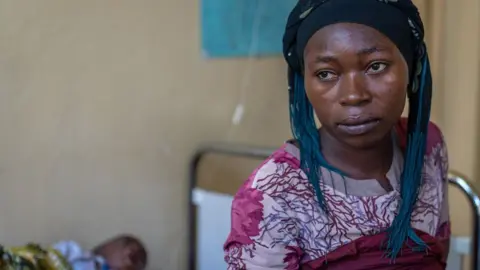 Julienne Mwinja sits on a hospital bed where her sick child lies - he is not in focus - in Lwiro clinic, DR Congo - November 2024