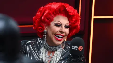 BBC A drag queen with permed red hair and painted red lips, wearing a black top and strings of pearls, smiles at the camera inside a radio studio.