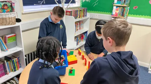 BBC Four primary school children stand around a table playing with lego.