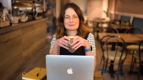 Linda Harrison A woman with long dark hair is sitting at a table in a cafe. She is wearing a black and white striped top with a black sleeveless top over it. She is holding a mug with both hands and looking at the camera over the top of her Apple laptop. She has a yellow notebook with a pen on top of it beside her. In the background are empty tables and chairs and a bar running along the left side of the cafe with a coffee machine on it.