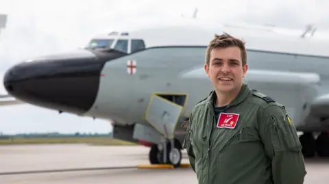 Sqd Ldr Dan Wilkes wearing his flying gear and standing beside an aircraft on a runway with field in the distance. He has his hands behind his back.