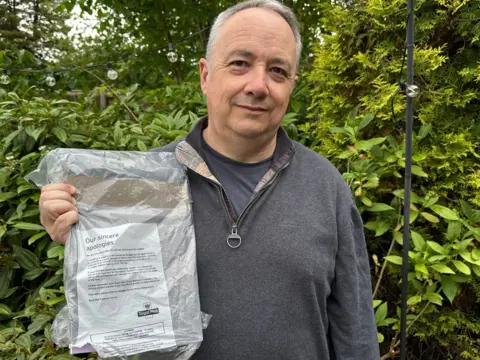 Paul Edwards A man in a grey quarter-zip holding a crumpled package from Royal Mail with an apology letter, stood in front of a green bush.