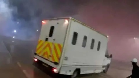 A screenshot of a police van seen on the Elbit Systems UK site. The van is a large white vehicle with small windows and a chevron pattern across the back. A figure wearing black with a white helmet can be seen in the lower right corner of the image holding a long unidentified object.