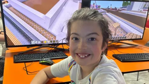 A young boy with long brown hair tied back in a ponytail wears a white t-shirt with colourful patterns on it. He sits in front of a large computer monitor, where a video game can be seen.