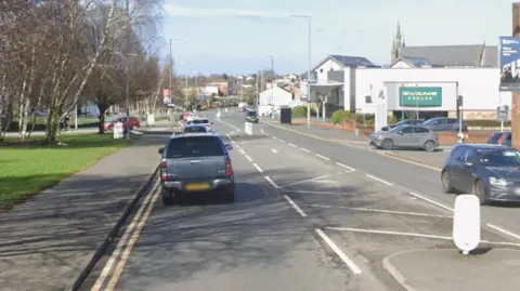 Google streetview of cars driving on North Road with trees on left and shops and properties on right