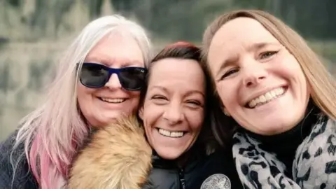 Three smiling faces of Suzy Angus, Shirley Ross and Emma Bryson are close together and looking at the camera. Suzy has light pink hair and glasses, Shirley has brown hair and a fur-collared jacket on, and Emma is closest to the camera with longer brown hair and a pink leopard scarf.