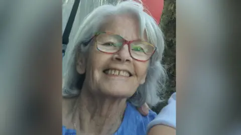 A close-up of the late Bernadette (Bernie) Cranley.  She has shoulder-lengthy grey hair and is wearing glasses with coloured rims. 