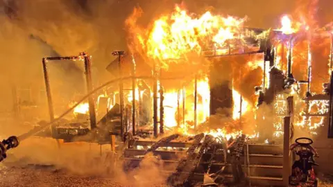 Essex Fire and Rescue Service A firefighter hoses down a large fire which has visibly gutted a structure.