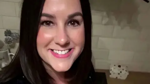 X Lucy Connolly with long straight dark hair smiling at the camera and wearing a black top. The photo is from a social media post and a table with a rock and artificial flowers is visible behind her.