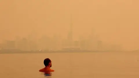 Toronto Star via Getty Images A man swimming in Lake Ontario on 15 July overlooking the Toronto skyline blanketed by a bright, orange haze from wildfire smoke. 