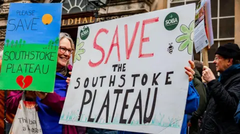 Jamie Bellinger Two large protest signs are held up, both reading "save the Southstoke Plateau". The signs are decorated, one with a green field, blue sky and a broken heart, and the other with green flowers and a bee. A smiling woman can be seen behind one of the signs.