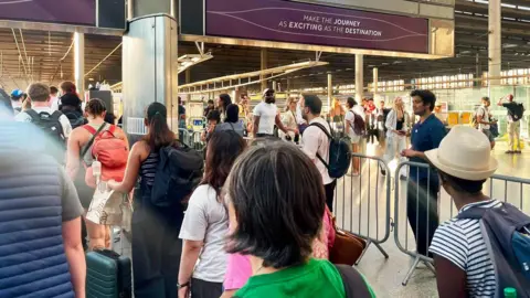 Nine people can be seen queuing at a train platform barrier at St Pancras station. There is a metal barrier beside them. At the other side of the barrier, people are walking in the opposite direction through another line of barriers. There is a departure board above the people which displays a purple sign reading "Make the journey as exciting as the destination"