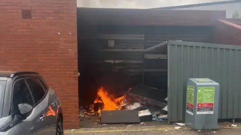 A fire in a bin store, with a metal structure to the right and a metal box. A car is to the left. 