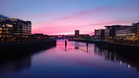 Davy McKenna The Clyde at sunset with buildings on either side and a bridge in the middle. The purple sky is reflected in the water