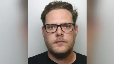 A mugshot of a bearded and bespectacled man in a black t-shirt. He has blond hair and looks straight at the camera.