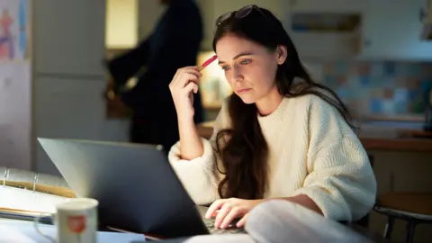 A young person is sitting at a table using a laptop in a softly lit kitchen. Glasses rest on the person's head while holding a pen near the forehead. Papers, a binder and a mug are visible in the foreground, with another blurred figure in the background.