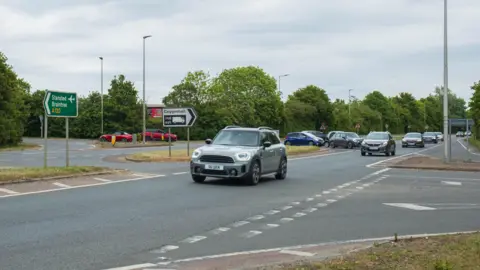 Paul Adams Cars driving along the single-lane A120 at the Colne Road junction. Several cars are queuing in an island on the middle of the road, waiting to join it.