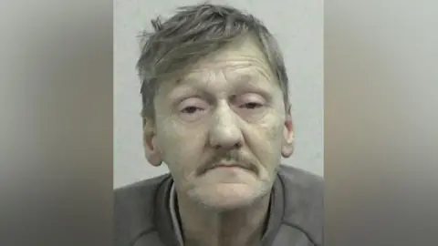 Mugshot of Howell. he has brown greying hair and a moustache. His skin is yellowing, his ears blotchy and he has a lot of wrinkles