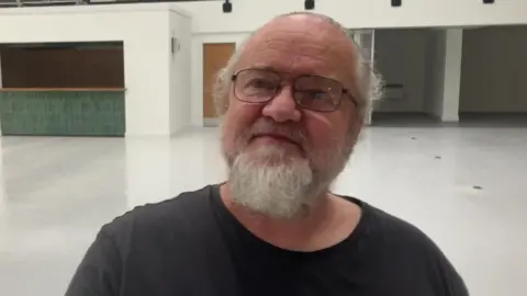 A man stands smiling looking up in a black tshirt. In the background, empty white stalls for market traders can be seen