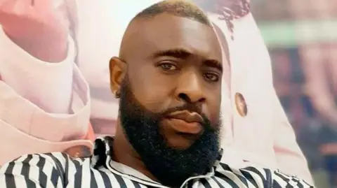 Macben Acianu, who has close-cropped black hair on top and sports a full beard, wearing a black and white striped shirt.