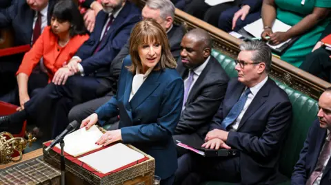 PA Chancellor, Rachel Reeves, at the despatch box in the house of commons delivering the budget. She wears a blue suit with red nail varnish, members of the cabinet, including the Prime Minister, Keir Starmer, sit behind her on green benches. 