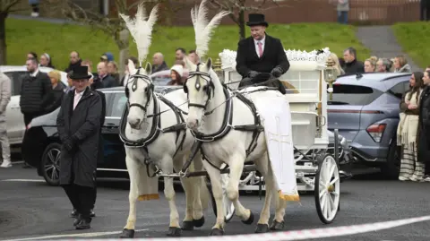 PA Media Two white horses wearing funeral gear pull a white carriage carrying Amy's coffin. Behind the horses a man holds the reigns, and beside them walks another funeral director, dressed in black, hands clasped solemnly. 
