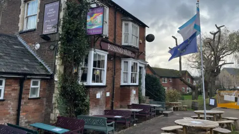 Tony Fisher/BBC Outside of a pub with tables and benches and a European Union flag flying with houses in the background.