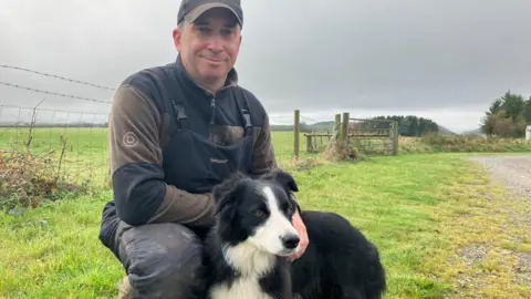 a man and a sheepdog by a fence on a farm