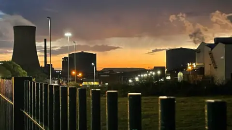 Buildings at Scunthorpe steelworks, including a cooling tower and tall chimney, in shadow at dusk, as lights shine against an orange and purple sky.