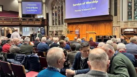 rows of men conversing with each other - there are about 40 people visible, in a church hall with large screens upon which the words Service of Worship/ Thursday 18 December 2025