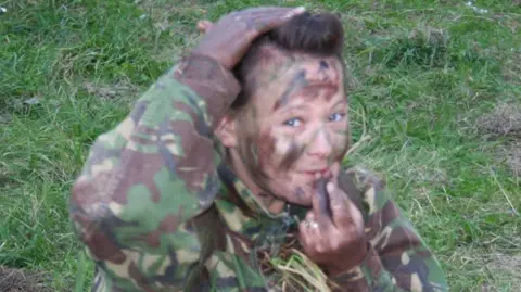 Family Handout A woman in camouflage gear and combat body paint smiling while sitting on a grassy bank