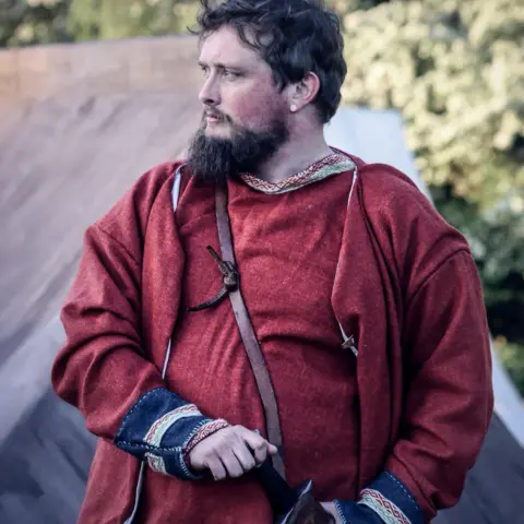 Handout A man in red Viking clothing looks to the left. He has dark hair and a beard.