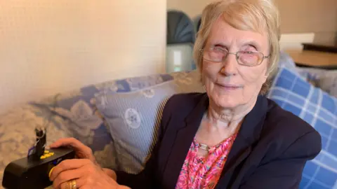 A woman with glasses is sitting on a sofa and  turning the knob of a radio. She is wearing a navy blazer and a pink colourful top underneath.