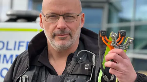 BBC DC Aaron Flint, wearing a black police uniform stands in front of a police car holding two catapults. One catapult is orange and another is silver coloured.
